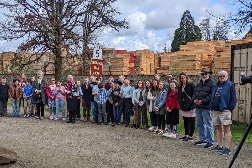 A crowd of 35 people stand along a park pathway watching the planting of the sapling
